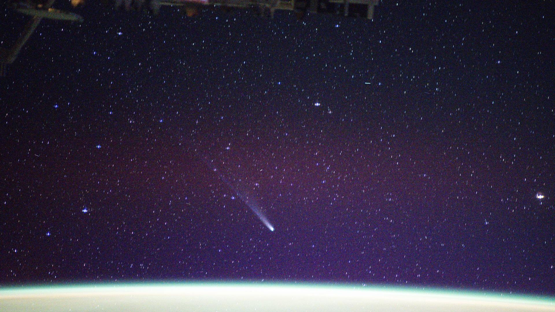 A streak of white is seen as comet Lemmon moves toward the bottom right of the image in a purple starry night sky. At the bottom are curved lights of white and green