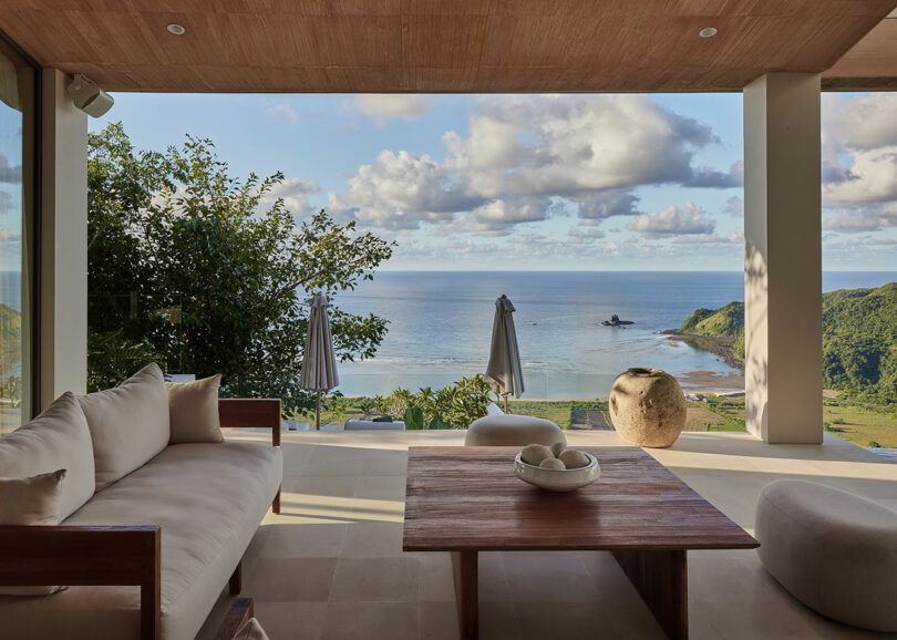 A modern outdoor patio with a wooden sofa set overlooks a coastal landscape, featuring ocean, beach, green hills, and partly cloudy sky.