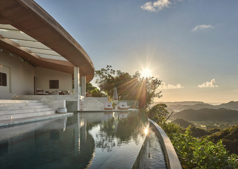 Modern hillside villa with an infinity pool overlooking a scenic valley at sunset, surrounded by lush greenery and distant mountains.
