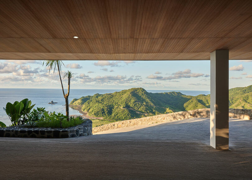 Open-air space with a wooden ceiling overlooks a scenic view of green hills, the ocean, and partly cloudy skies. A small garden with tropical plants is on the left.