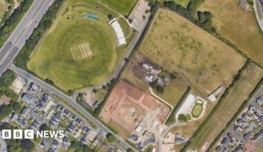 An aerial view of two fields which were formerly a golf driving range, surrounded by housing estates, roads and trees.