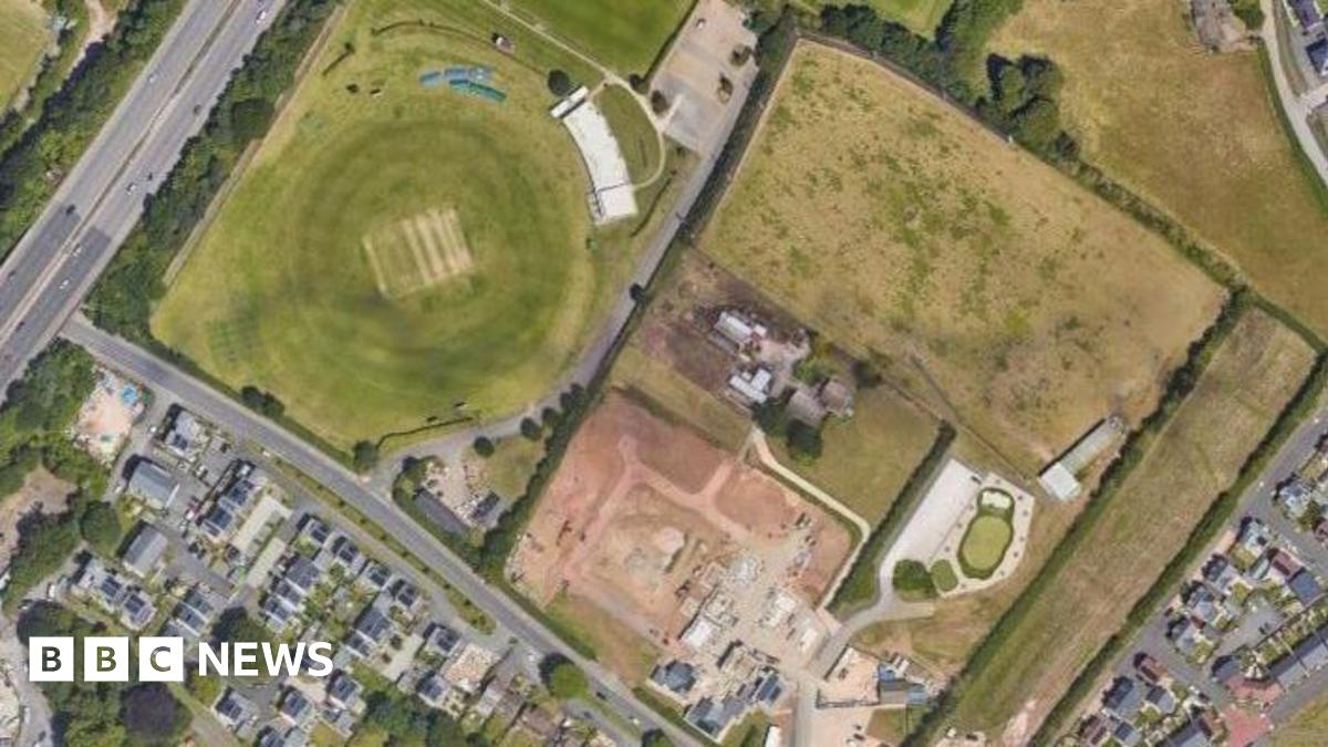 An aerial view of two fields which were formerly a golf driving range, surrounded by housing estates, roads and trees.