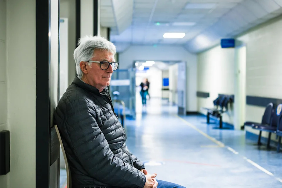 Portrait of a senior man in his 70s sitting and waiting in a hospital corridor. Focus on the man with a worried and anxious expression on his face, while the background of the hospital is defocused.