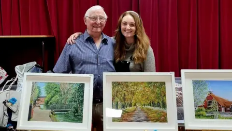 Robert Rose Robert Rose and granddaughter Joanna Menzies standing behind three white-framed prints of his paintings - a bridge, path through autumn leaves and a church