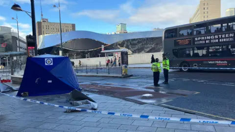 A blue police tent is on a pavement and cordoned off by blue and white police tape. There are two police officers standing inside the taped area wearing black hats and yellow high-visibility yellow jackets. There is a bus driving past in the background.