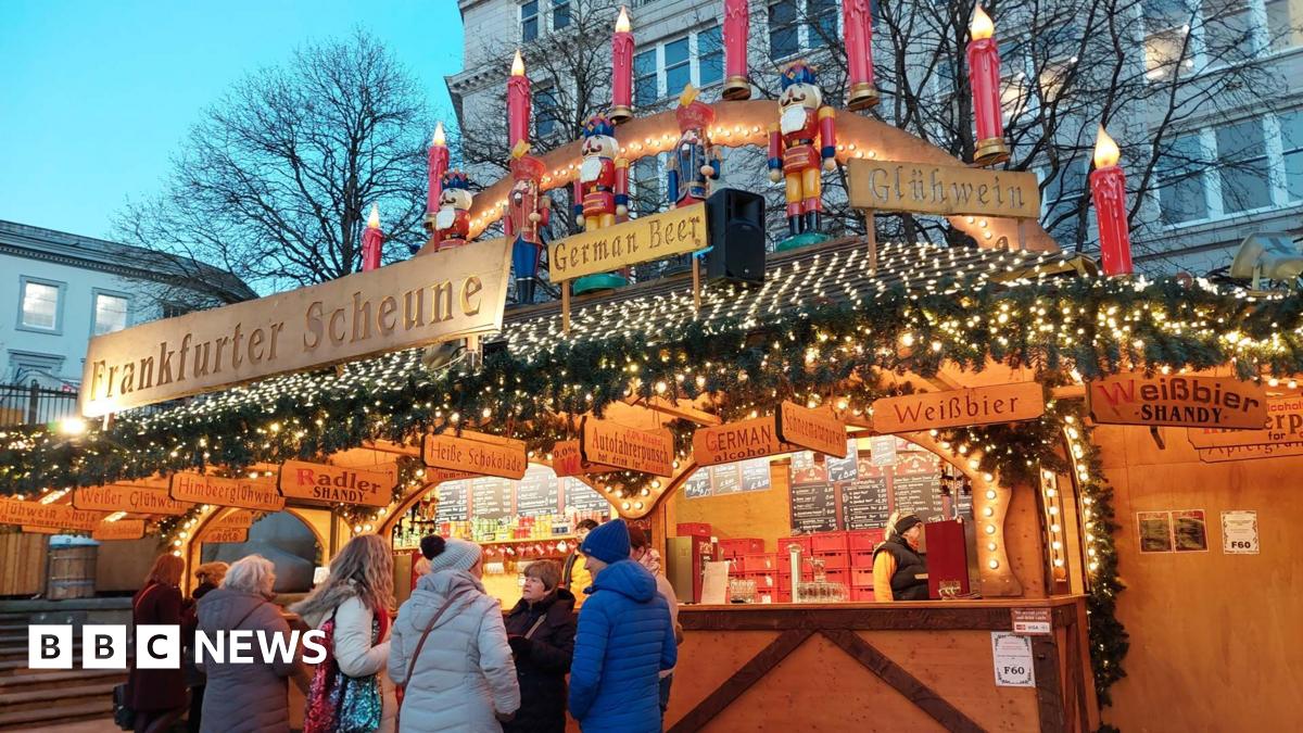 A stall selling churros at a Christmas market. The wooden stall is adorned with fake Christmas tree sprigs, golden fairylights, signs saying "churros". A man stands behind the counter at the stall, serving a woman in a long winter coat