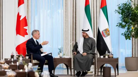 Reuters Sheikh Mohamed bin Zayed Al Nahyan, President of the United Arab Emirates, meets with Canadian Prime Minister Mark Carney at Al Shati Palace in Abu Dhabi, United Arab Emirates, November 20, 2025. The two are seated across from each other. Carney is wearing a black suit, while the Al Nahyan is wearing a traditional Emirati thobe and white gutra. Behind them are the UAE and Canadian flags. 