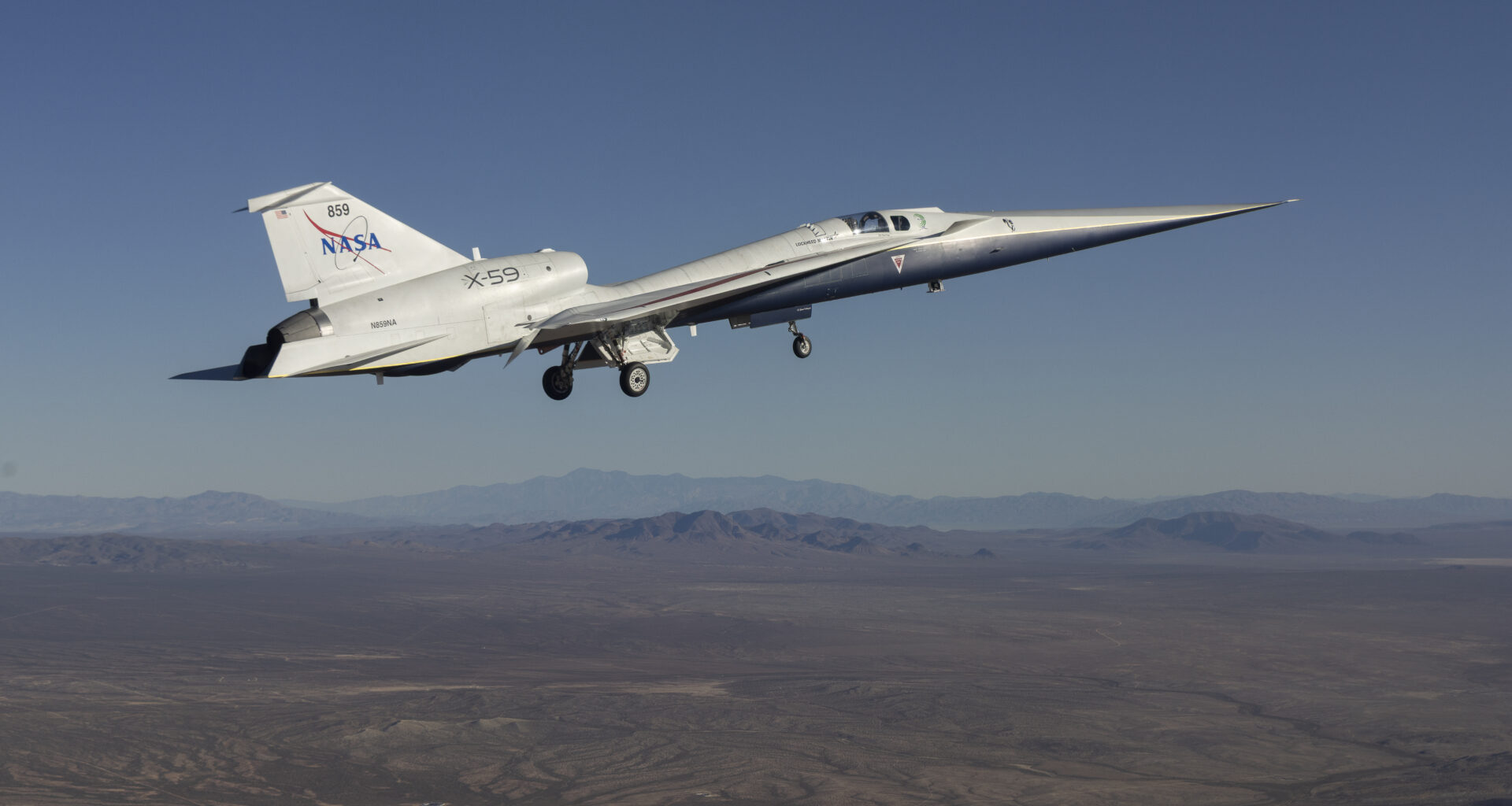 The X-59 quiet supersonic research aircraft flies above California. The plane has a distinctive shape with a long, sharply pointed nose. The nose is silver, while the rest of the body looks white. The words "NASA" and "X-59" are on the body of the aircraft. In the background, we can see brown earth below the plane as well as hazy mountains in the distance.