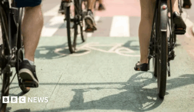 A number of bicycles are locked to two lines of bicycle racks on a small patch of concrete. To the right side, a small road leads to a white metal gate, and there is wooden fence behind and alongside the bikes. Behind that is a green racecourse with some trees, and houses in the background.