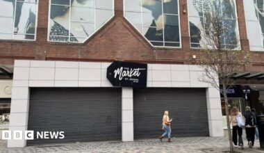 A woman walks past the external entrance to St Johns Market in Liverpool which has two black shutters down. A sign reads The Market, St Johns in white writing on a black background.