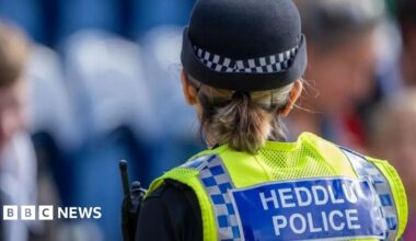 A female police officer wearing a hat and uniform faces away from the camera. Her uniform reads: HEDDLU POLICE