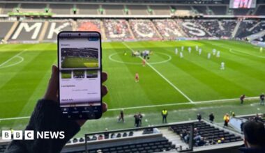 Image of someone sitting in a football stadium holding up a mobile phone, with images of a football pitch on it.  In the background can be seen a number of empty stadium seats, a football pitch, football players and officials.