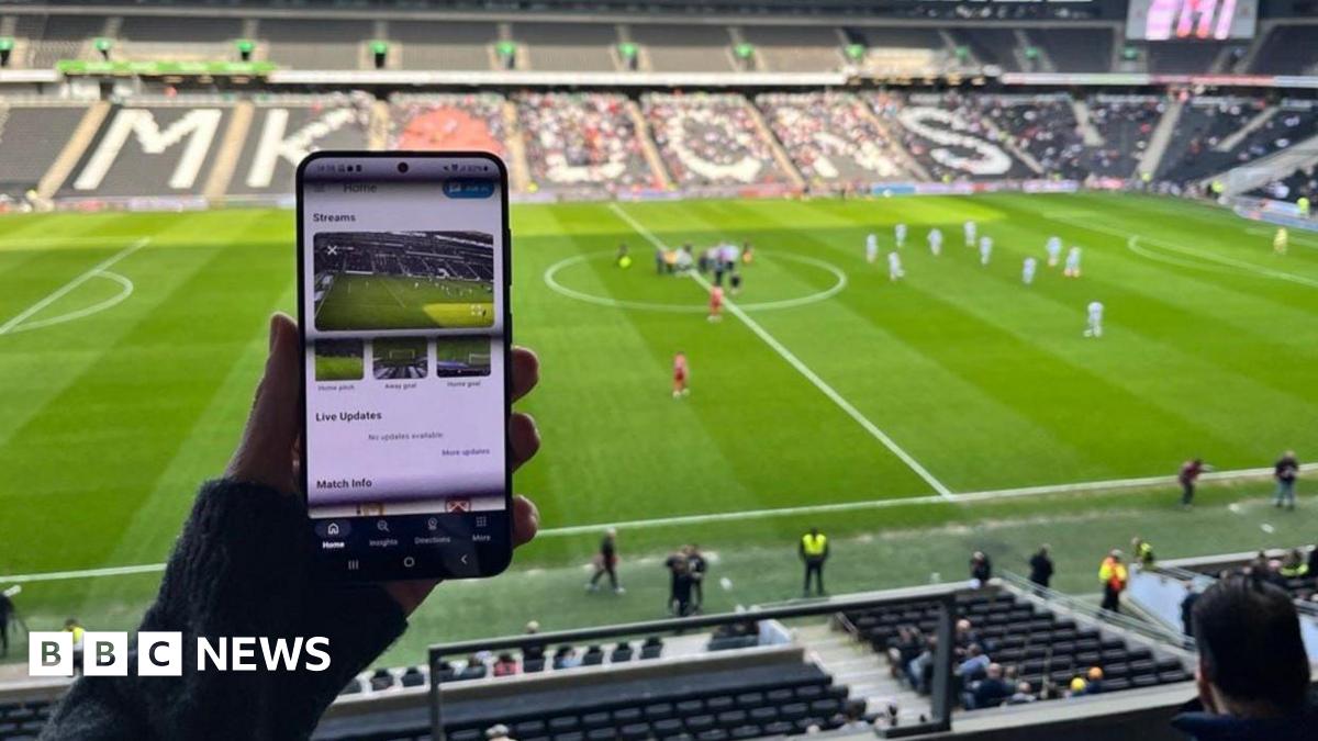 Image of someone sitting in a football stadium holding up a mobile phone, with images of a football pitch on it.  In the background can be seen a number of empty stadium seats, a football pitch, football players and officials.