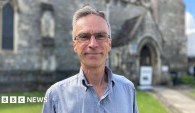 Dr Murrison smiles at the camera - he is wearing glasses and a blue shirt. The background is blurred, but it is a sunny day in front of a church