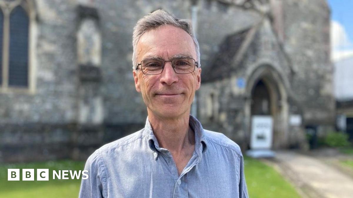 Dr Murrison smiles at the camera - he is wearing glasses and a blue shirt. The background is blurred, but it is a sunny day in front of a church