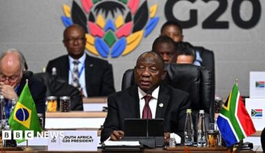 South African President Cyril Ramaphosa seated (right) in black suit and white shirt and microphone and a tablet. In front of him on the table is a South African flag poster written "South Africa G20 President'