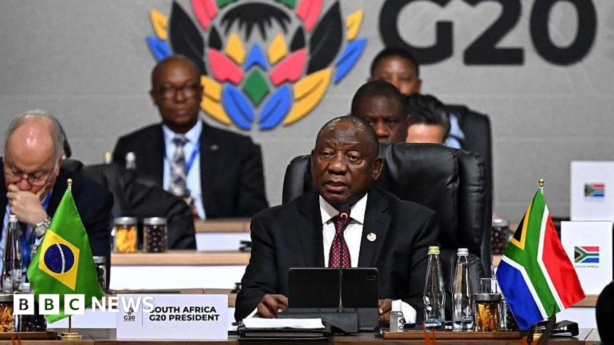 South African President Cyril Ramaphosa seated (right) in black suit and white shirt and microphone and a tablet. In front of him on the table is a South African flag poster written "South Africa G20 President'