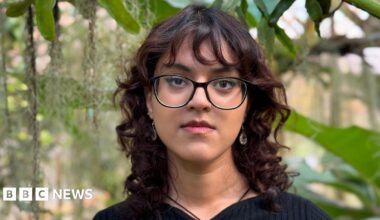 Benika has curly red-brown shoulder-length hair and black glasses. She is wearing a black jumper, and there are green leaves behind her.