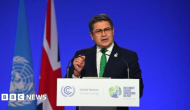 Honduras President Juan Orlando Hernández speaking at a podium, wearing a black suit and a green dotted tie. Behind him is a blue backdrop with a UN flag and a UK flag to his left. He has short hair and wears square-framed eye glasses.