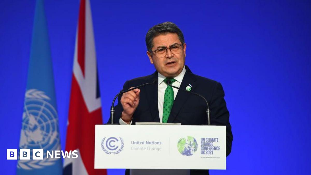 Honduras President Juan Orlando Hernández speaking at a podium, wearing a black suit and a green dotted tie. Behind him is a blue backdrop with a UN flag and a UK flag to his left. He has short hair and wears square-framed eye glasses.