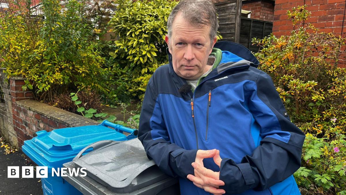 Stuart Atkinson has short grey hair and is wearing a blue waterproof jacket. He is leaning against a grey wheelie bin while looking at the camera.
