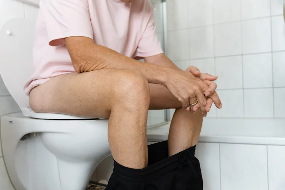 Person sitting on a toilet in a bathroom, wearing a light shirt, with their hands clasped and looking thoughtful