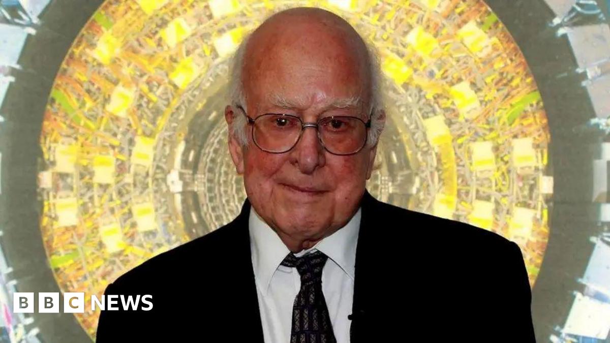 Prof Higgs is looking at the camera. He is bald apart from some white tufts of hair at the sides of his head. He is wearing glasses and a dark suit and tie.There is a bright yellow and green circular background