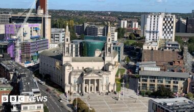 Leeds Civic Hall, a white stone building with a blue sky behind. To the right is a golden clock attached to the building and a golden owl on a pillar. There are pedestrians walking on the pavements outside.