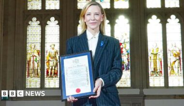 Cate Blanchett, a woman with long, blonde hair, wears a grey suit and hold up a framed certificate. She stands in front of a row of stained glass windows with light pouring in.