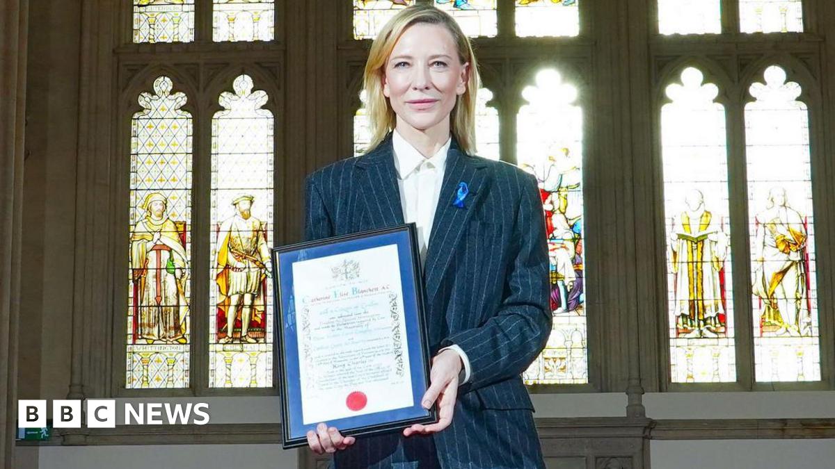 Cate Blanchett, a woman with long, blonde hair, wears a grey suit and hold up a framed certificate. She stands in front of a row of stained glass windows with light pouring in.