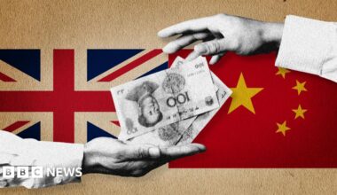 Sir Jeremy Fleming is dressed in a dark navy suit jacket with structured shoulders, a crisp white dress shirt, and a pale blue textured tie. He is seated in a dimly lit indoor setting with soft shadows and a dark, neutral background. The lighting highlights the suit fabric and tie texture, suggesting a formal or professional environment.