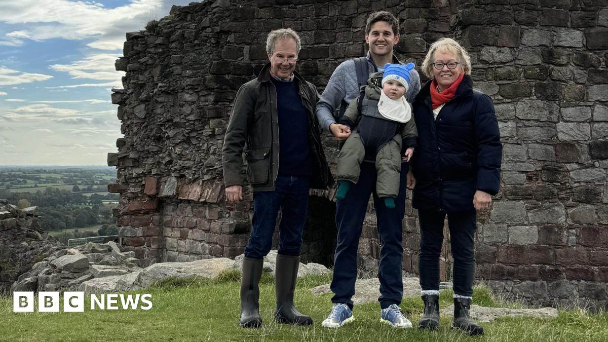 A family group standing outdoors in front of the remains of an old stone wall, part of an historic ruin or castle. The people are dressed in outdoor clothing, including jackets and boots, and the man in the centre is carrying a small child. Behind the group there is a scenic view of green fields stretching into the distance under a partly cloudy sky.