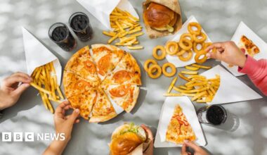 Looking down on a table full of plates of pizza, burgers in buns, onion rings, french fries and cola drinks, which are all examples of ultra-processed foods. Hands are reaching out and picking up the different foods.