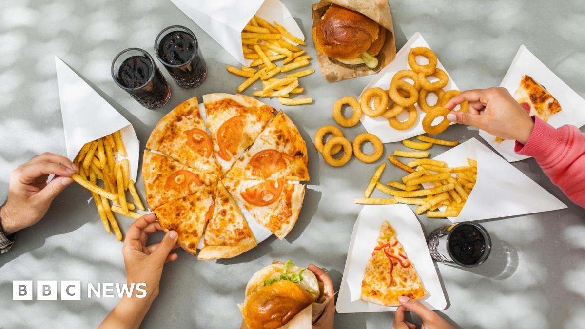 Looking down on a table full of plates of pizza, burgers in buns, onion rings, french fries and cola drinks, which are all examples of ultra-processed foods. Hands are reaching out and picking up the different foods.