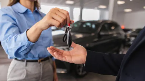 Getty Images Saleswoman handing over car keys to businessman, finalising a successful vehicle purchase in a sleek, modern car dealership showroom.