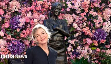 Renée Zellweger smiles while wearing a black dress and posing next to a statue of Bridget Jones next to a sign that says 'Bridget Jones' against a backdrop of pink and purple flowers lining the wall, in Leicester Square in London on Monday.