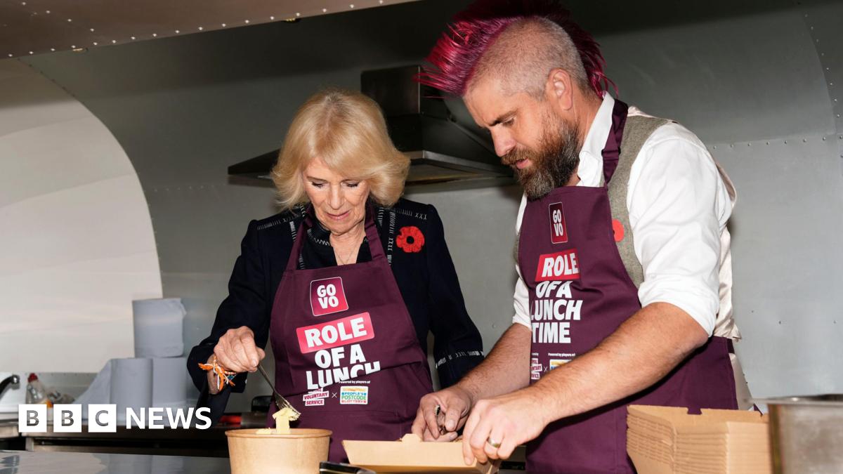 Queen Camilla, wearing a navy blue dress and a poppy with a burgundy apron, is seen talking to Ben Newman who has a pink mohawk and wearing a white shirt and grey waistcoat plus a burgundy apron. They are standing outdoors, with other people visible in the background.