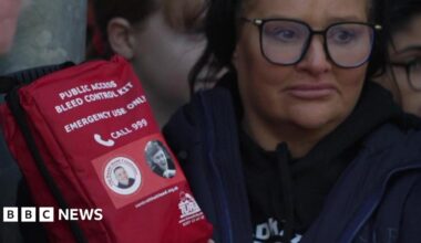 A woman wearing black-rimmed glasses holds a red package marked "bleed control" kit and which also bears a picture of Harvey Willgoose