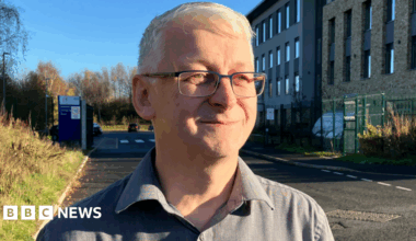 Dr Robert Westgate, with short white hair, glasses, and wearing a grey shirt, looks to the right of the camera, as he stands on a road outside a large building on the right, with rectangular windows. There is a car park entrance behind him.