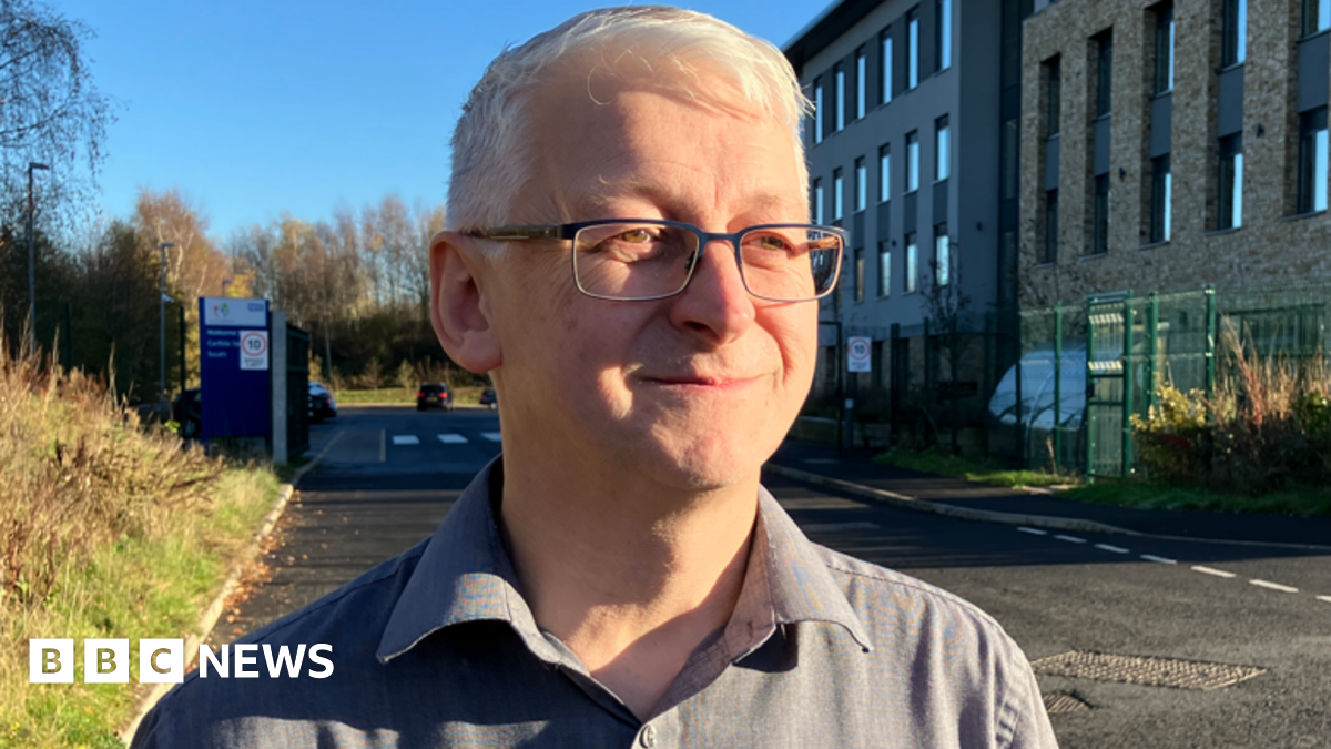 Dr Robert Westgate, with short white hair, glasses, and wearing a grey shirt, looks to the right of the camera, as he stands on a road outside a large building on the right, with rectangular windows. There is a car park entrance behind him.