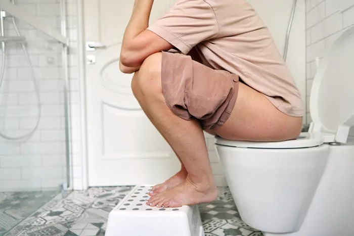 Person demonstrating a squatting position on a toilet using a footstool, promoting ergonomic posture for easier bowel movements