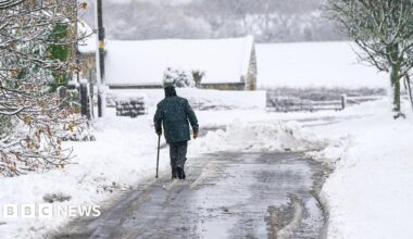 Man walks on a road partially cleared of snow, with a covering of snow on trees in the foreground and a barn and fields in the background