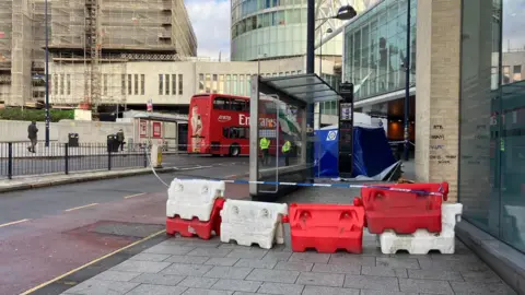 Red and white blocks are blocking off a pavement and there is blue and white police tape. A silver bus stop is behind the tape and there is a blue police tent on the pavement beyond it.