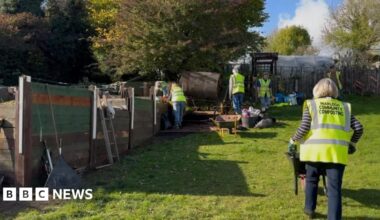 Compost mentor, Ben Bryant, who has auburn, tied back hair and matching beard, wearing a khaki fisherman's cotton tunic with pockets, standing in front of a compost tumbling machine with two volunteers sitting in the background.