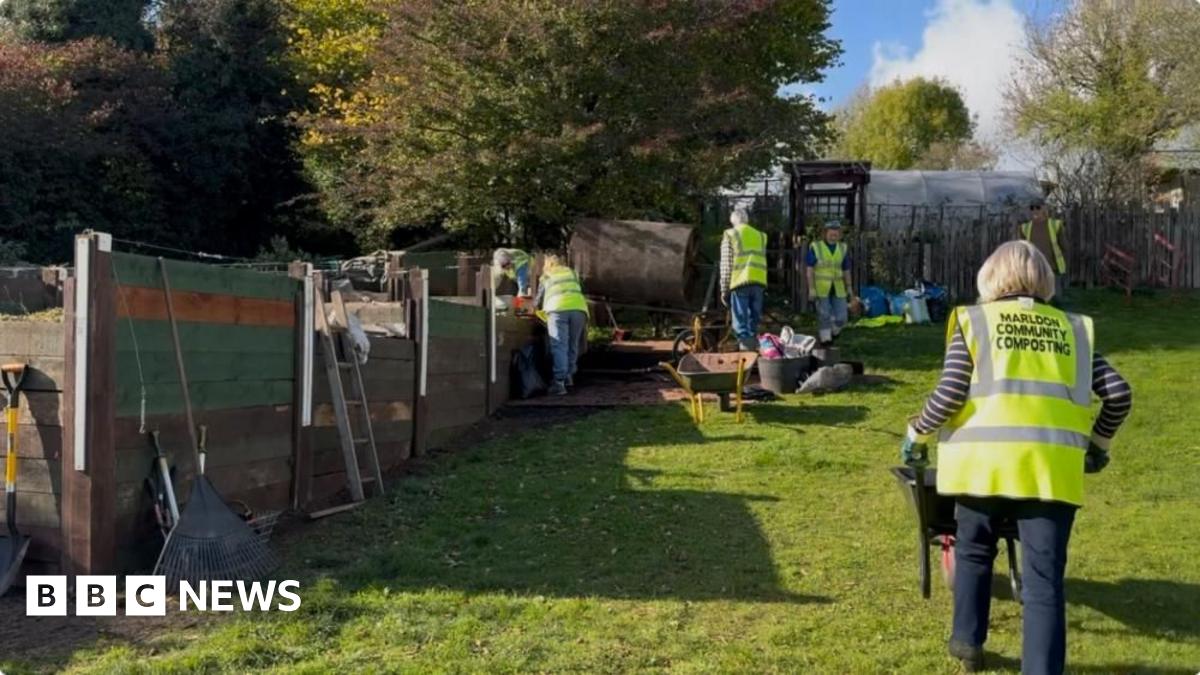 Compost mentor, Ben Bryant, who has auburn, tied back hair and matching beard, wearing a khaki fisherman's cotton tunic with pockets, standing in front of a compost tumbling machine with two volunteers sitting in the background.