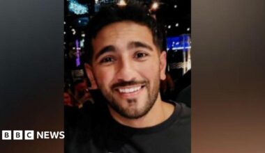 An image of a man with short dark hair, a moustache and beard smiling at the camera. He is wearing a black jumper and appears to be standing in a nightclub.