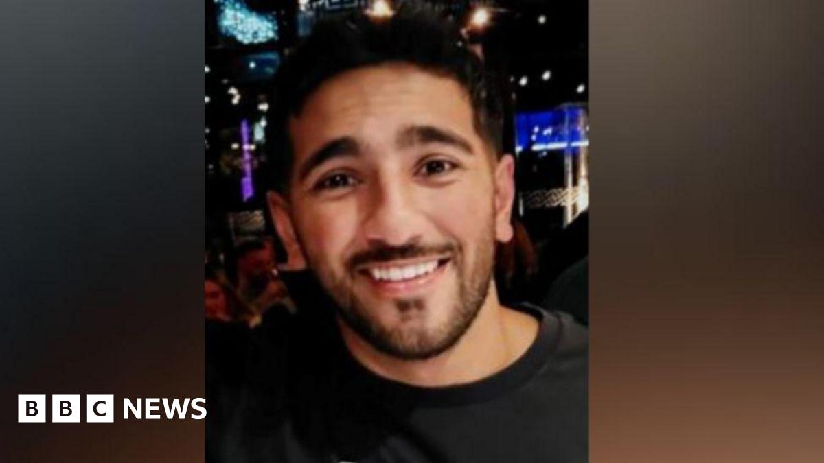 An image of a man with short dark hair, a moustache and beard smiling at the camera. He is wearing a black jumper and appears to be standing in a nightclub.