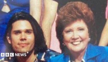 Cilla Black and Zinc Grantham sitting next to each other and smiling at the camera following a Blind Date TV show in 1997. Mr Grantham has shoulder-length black hair and is wearing a multi-coloured shirt. Cilla Black is wearing a royal blue jacket with a blue and white shirt. She has short wavy red hair.