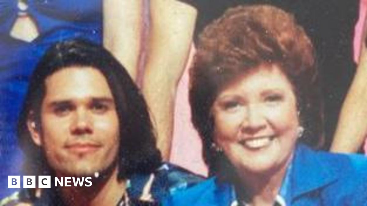 Cilla Black and Zinc Grantham sitting next to each other and smiling at the camera following a Blind Date TV show in 1997. Mr Grantham has shoulder-length black hair and is wearing a multi-coloured shirt. Cilla Black is wearing a royal blue jacket with a blue and white shirt. She has short wavy red hair.