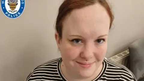 West Midlands Police A woman wearing a black and white-striped top sits down and looks up at the camera with a smile. A white wall and radiator are behind her.
